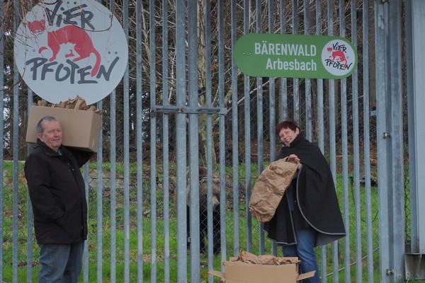 Zwei bÄRENWALD-Unterstützer: ein Mann und eine Frau stehen mit den gespendeten Futtersäcken vor einem Gittertor. Hinter dem Gittertor schaut Bär Erich hervor