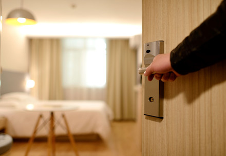 eine Hand öffnet eine Hotelzimmertür, man blickt in das Zimmer, man sieht ein Bett, ein Tisch und ein Fenster durch den das Licht scheint